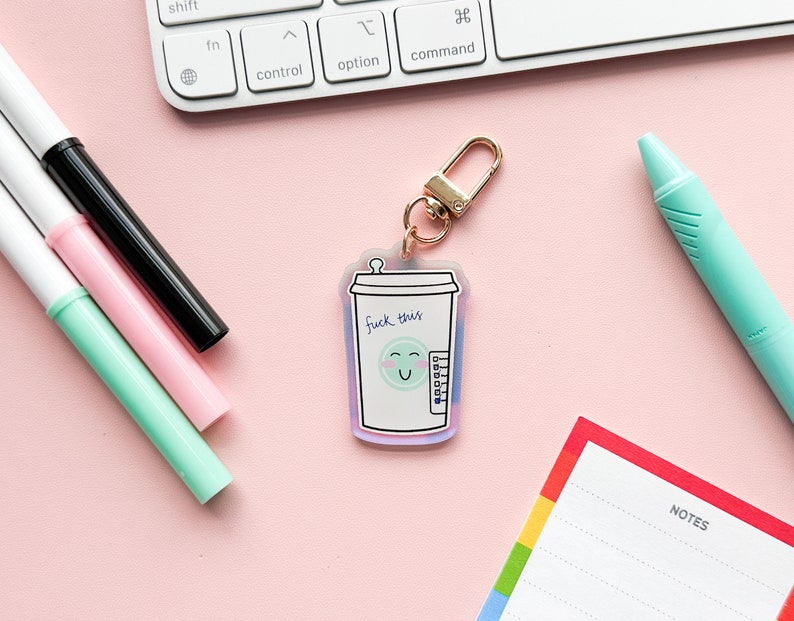 May include: A gold keychain with a clear acrylic charm featuring a cartoon coffee cup with a smiling face and the text "F*ck this".