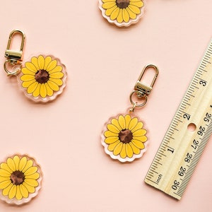 Peut inclure: Quatre porte-cl&eacute;s en acrylique transparent avec un fermoir en m&eacute;tal dor&eacute;. Chaque porte-cl&eacute;s pr&eacute;sente un tournesol jaune avec un centre brun. Les porte-cl&eacute;s sont dispos&eacute;s sur une surface rose avec une r&egrave;gle en bois.