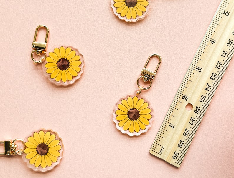Peut inclure: Quatre porte-cl&eacute;s en acrylique transparent avec un fermoir en m&eacute;tal dor&eacute;. Chaque porte-cl&eacute;s pr&eacute;sente un tournesol jaune avec un centre brun. Les porte-cl&eacute;s sont dispos&eacute;s sur une surface rose avec une r&egrave;gle en bois.