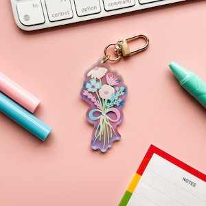 May include: A pink and blue floral bouquet keychain with a gold metal ring. The keychain is made of clear acrylic and features a colorful floral design with a pink ribbon.