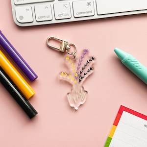 May include: A clear acrylic keychain with a floral design. The design features purple, yellow, and black flowers with a gold metal key ring.