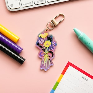 Może przedstawiać: Brelok w kształcie kolorowego bukietu kwiatów ze złotym metalowym kółkiem. Bukiet wykonany jest z akrylu i zawiera różne kwiaty w odcieniach żółtego, fioletowego i różowego. Brelok to świetny sposób na dodanie odrobiny fantazji do kluczy lub torebki.