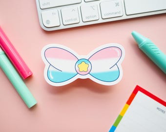 May include: A bow-shaped sticker with a design featuring the colors of the transgender flag: pink, white, and light blue. A yellow star is in the center. The sticker is on a pink surface, with pens, a keyboard, and a notepad.