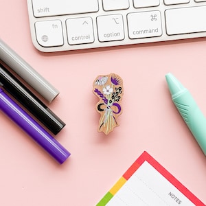 May include: A wooden flower bouquet pin with purple, white and black flowers. The pin is on a pink surface with a keyboard, pens and a notepad.