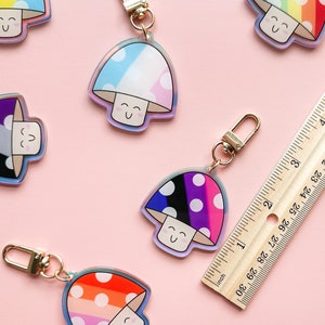 Puede incluir: Cinco llaveros coloridos con forma de hongo con lunares blancos y caras sonrientes. Cada llavero presenta una combinaci&oacute;n de colores diferente, incluyendo arco&iacute;ris, azul y rosa, negro y blanco, y gris y blanco.
