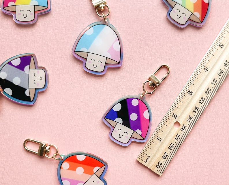 Puede incluir: Cinco llaveros coloridos con forma de hongo con lunares blancos y caras sonrientes. Cada llavero presenta una combinaci&oacute;n de colores diferente, incluyendo arco&iacute;ris, azul y rosa, negro y blanco, y gris y blanco.