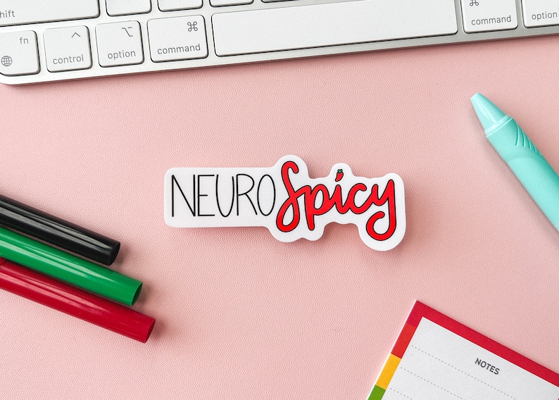Puede incluir: Una pegatina blanca con el texto "NEUROspicy" en letras negras y rojas. La pegatina est&aacute; sobre una superficie rosa con un teclado, un bloc de notas y marcadores.