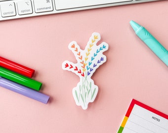 Può includere: Un adesivo a forma di bouquet di fiori di lavanda dai colori dell'arcobaleno. L'adesivo è su una superficie rosa con una tastiera, una penna verde e tre pennarelli sullo sfondo.