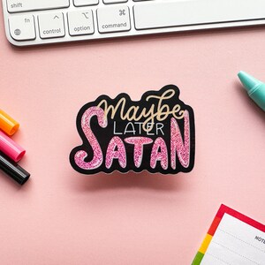 May include: A pink glitter sticker with the text "Maybe Later Satan" on a black background. The sticker is on a pink surface with a keyboard, pens, and a notepad in the background.