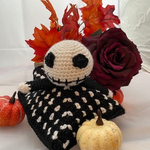 May include: A crocheted Halloween-themed doll with a black and white skeleton pattern. The doll is sitting on a blanket with two small pumpkins beside it. The doll is surrounded by fall foliage, including a red rose.