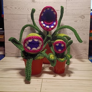 May include: Crocheted plant sculptures in orange pots. The plants have large, open mouths with white teeth, pink and purple interiors, and green stems and leaves. The plants are arranged on a wooden surface.