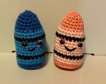 May include: Two crocheted crayon toys with smiling faces and googly eyes. One crayon is blue and the other is pink.