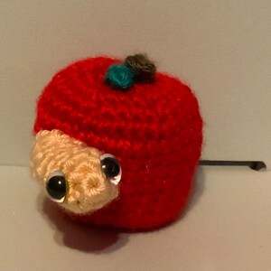 May include: A crocheted red apple with a white worm peeking out and a blue and brown flower on top.