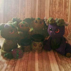 May include: A collection of four crocheted creatures with large, glossy eyes and textured yarn bodies. Three are olive green, and one is purple with green accents. Two small, clear plastic jars with black lids are positioned among the figures.