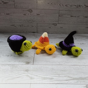 May include: Three handmade crochet turtles in Halloween colors. One is black with a purple top, another is yellow with a candy corn shell, and the third is black with a witch hat. The turtles have green bodies and black eyes.
