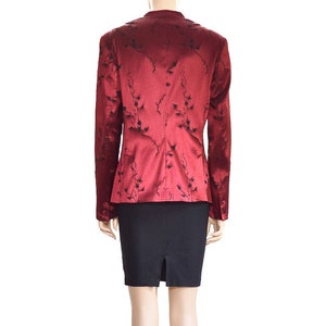 May include: A red blazer with a black floral pattern. The blazer has a fitted silhouette and long sleeves. The model is wearing a black skirt and black heels.