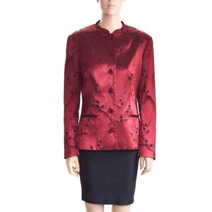May include: A red satin blazer with a black floral pattern and a mandarin collar. The blazer has a single button closure and two pockets. The model is wearing a black skirt and black sandals.