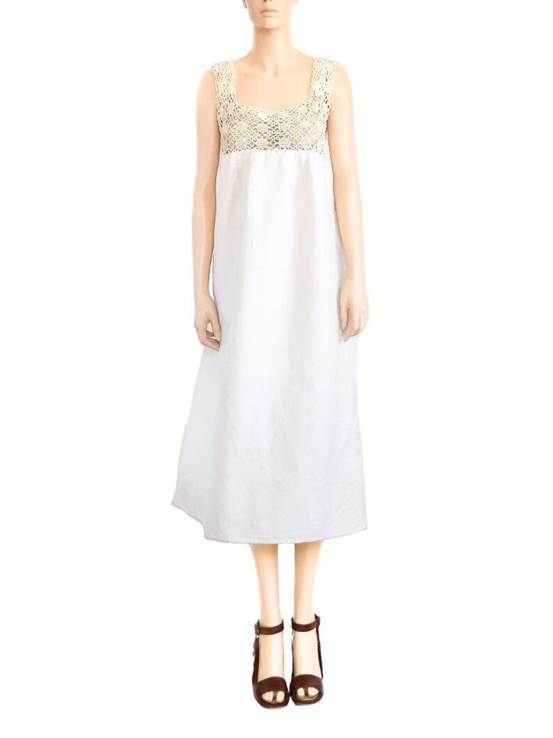 May include: A white linen dress with a crocheted bodice and a simple, flowy silhouette. The dress has a square neckline and sleeveless design. The model is wearing brown heeled sandals.