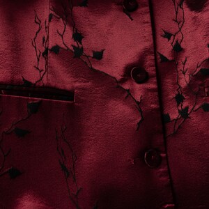 May include: A close-up of a burgundy fabric with a black floral pattern. The fabric has a satin-like sheen and features several buttons.
