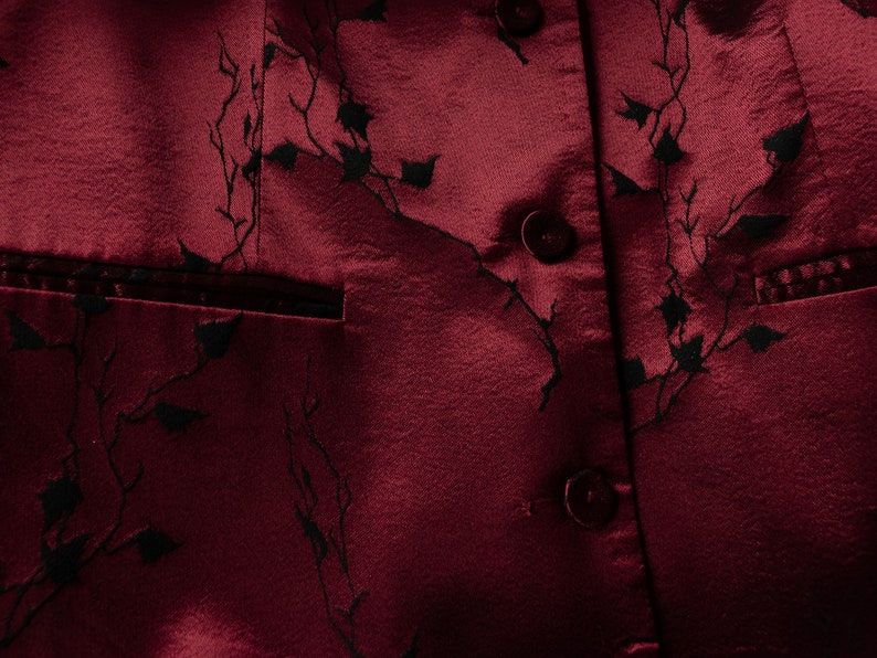 May include: A close-up of a burgundy fabric with a black floral pattern. The fabric has a satin-like sheen and features several buttons.