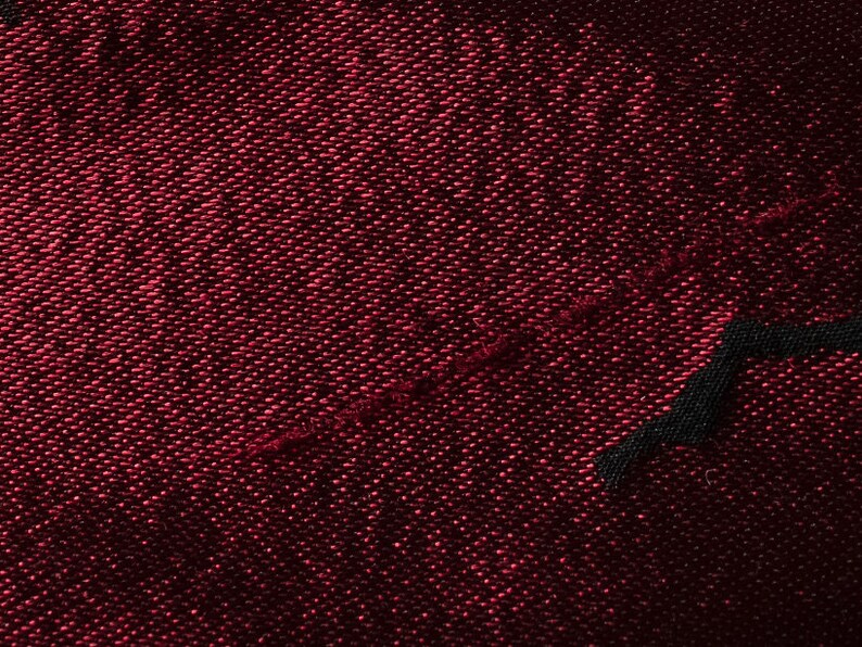 May include: Close-up of a dark red fabric with a textured weave. The fabric has a subtle sheen and a diagonal seam.