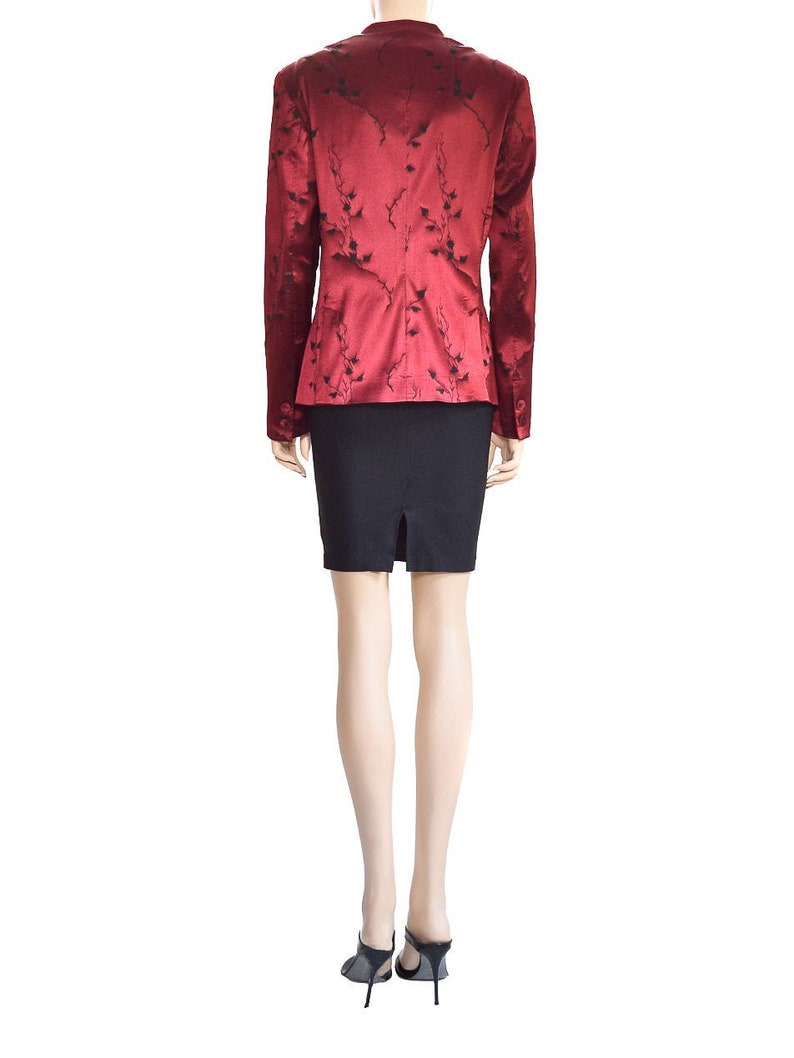 May include: A red blazer with a black floral pattern. The blazer has a fitted silhouette and long sleeves. The model is wearing a black skirt and black heels.