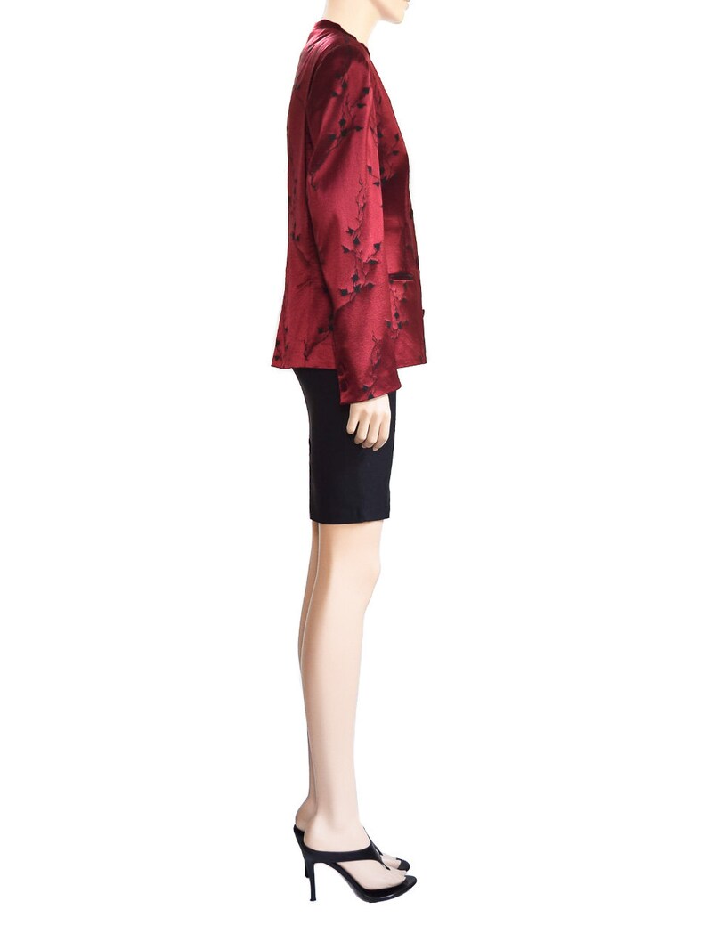 May include: A red satin blazer with a black floral pattern. The blazer has a mandarin collar and long sleeves. The model is wearing a black skirt and black heels.