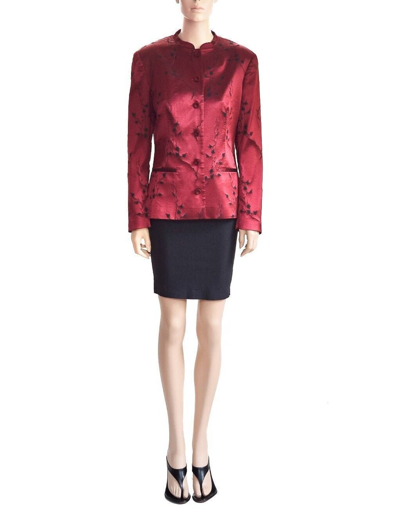 May include: A red satin blazer with a black floral pattern and a mandarin collar. The blazer has a single button closure and two pockets. The model is wearing a black skirt and black sandals.