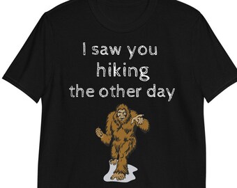 May include: Black t-shirt with the text "I saw you hiking the other day" above a cartoon illustration of Bigfoot pointing. The Bigfoot is brown and standing on a patch of white.