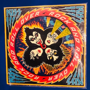 Puede incluir: Una portada de álbum cuadrada con la banda KISS. La portada tiene un fondo negro con las caras de los miembros de la banda en un círculo. Las palabras "KISS" y "ROCK AND ROLL OVER" están en un diseño rojo y azul.