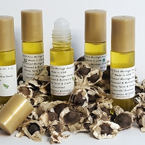 May include: Five small glass bottles of moringa seed oil, with gold lids and labels, are arranged on a bed of moringa seeds. The labels indicate that the oil is 100% pure, made in the USA, and processed and bottled by Paisley Farm & Crafts. The bottles are unscented and have an expiration date of 9/2020.