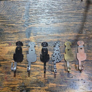 May include: Six metal wall hooks shaped like sitting dogs, arranged in a row. The hooks feature various finishes including solid black, matte gray, dark metallic, a spotted dalmatian pattern, brushed gold, and a copper-toned finish.