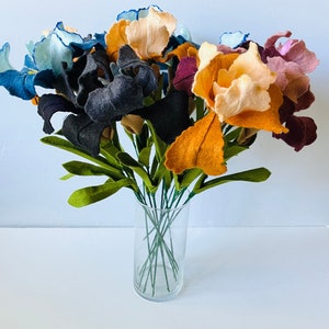 May include: A bouquet of felt iris flowers in a clear glass vase. The flowers are in various colors, including blue, black, orange, and pink. The green leaves and stems add contrast to the colorful blooms.