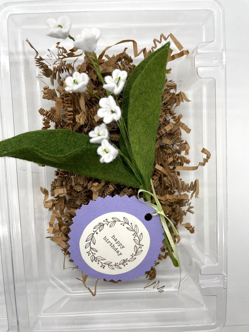 Peut inclure: Un petit bouquet de fleurs de muguet en feutre blanc avec des feuilles en feutre vert dans un r&eacute;cipient en plastique transparent avec du papier d&eacute;chiquet&eacute; brun. Une &eacute;tiquette violette avec le texte "joyeux anniversaire" est attach&eacute;e au bouquet.