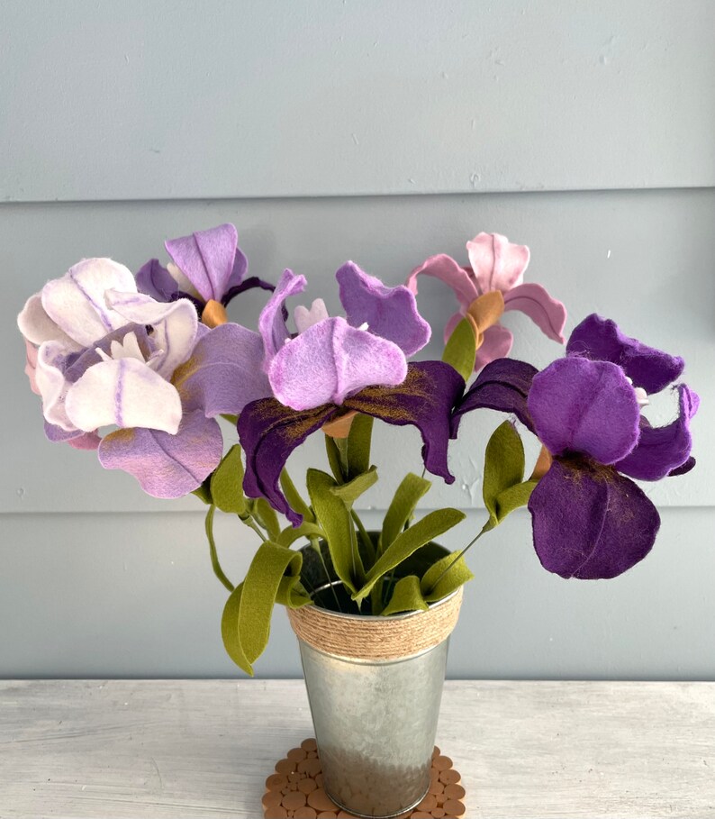 May include: A bouquet of purple and white felt iris flowers in a galvanized metal vase with a burlap ribbon around the top. The vase is sitting on a wooden surface.