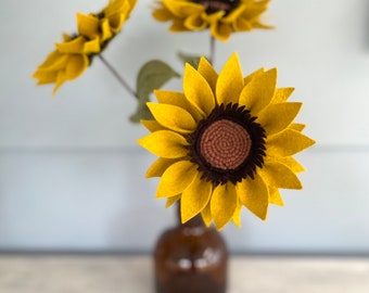 May include: Three felt sunflowers in a brown glass vase. The sunflowers are yellow with brown centers and dark brown petals. The vase is a simple, round shape.