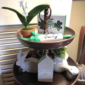 Peut inclure: Un plateau &agrave; deux &eacute;tages avec une base en m&eacute;tal brun et une figurine de lapin en c&eacute;ramique blanche. Le niveau sup&eacute;rieur pr&eacute;sente un rouleau &agrave; p&acirc;tisserie vert avec le mot &laquo;&nbsp;lucky&nbsp;&raquo; &eacute;crit dessus, une petite plante en pot avec des fleurs blanches et une carte verte et blanche avec le texte &laquo;&nbsp;Luck & Blessed&nbsp;&raquo;. Le niveau inf&eacute;rieur pr&eacute;sente une pancarte en bois gris avec le texte &laquo;&nbsp;Hello Spring&nbsp;&raquo;, une figurine d'oiseau en c&eacute;ramique blanche et une figurine de lapin en bois gris avec le texte &laquo;&nbsp;bunny kisses & easter wishes&nbsp;&raquo;.