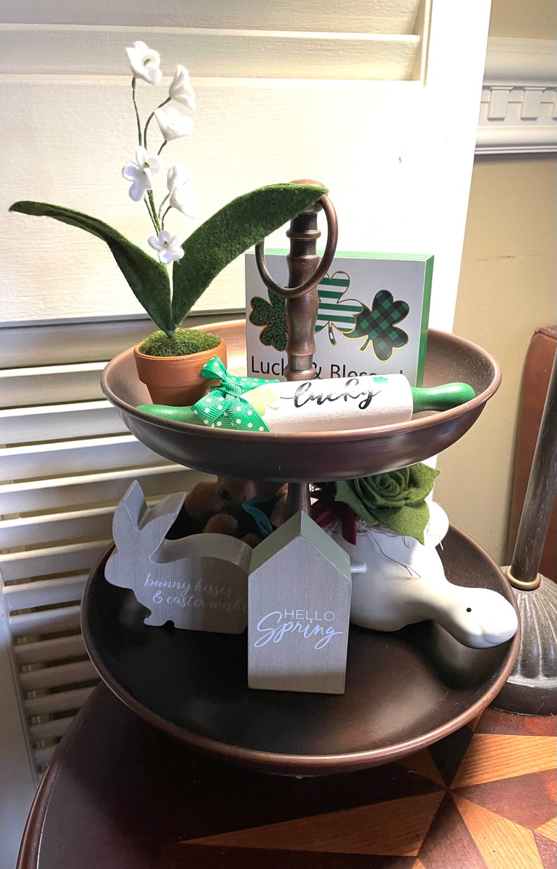 Peut inclure: Un plateau &agrave; deux &eacute;tages avec une base en m&eacute;tal brun et une figurine de lapin en c&eacute;ramique blanche. Le niveau sup&eacute;rieur pr&eacute;sente un rouleau &agrave; p&acirc;tisserie vert avec le mot &laquo;&nbsp;lucky&nbsp;&raquo; &eacute;crit dessus, une petite plante en pot avec des fleurs blanches et une carte verte et blanche avec le texte &laquo;&nbsp;Luck & Blessed&nbsp;&raquo;. Le niveau inf&eacute;rieur pr&eacute;sente une pancarte en bois gris avec le texte &laquo;&nbsp;Hello Spring&nbsp;&raquo;, une figurine d'oiseau en c&eacute;ramique blanche et une figurine de lapin en bois gris avec le texte &laquo;&nbsp;bunny kisses & easter wishes&nbsp;&raquo;.