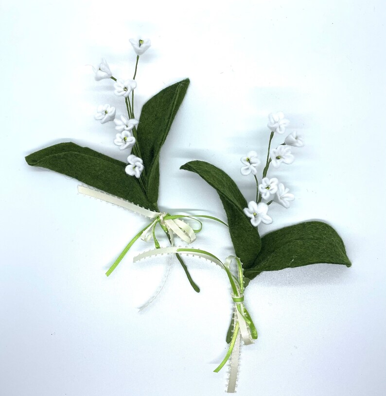 Peut inclure: Deux brins de muguet en feutre blanc avec des feuilles en feutre vert et des n&oelig;uds de ruban blanc et vert.