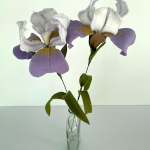 May include: Two purple and white felt iris flowers with yellow centers in a clear glass vase with a textured surface.