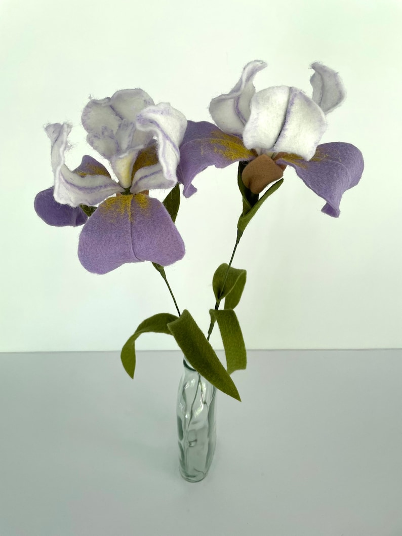 May include: Two purple and white felt iris flowers with yellow centers in a clear glass vase with a textured surface.