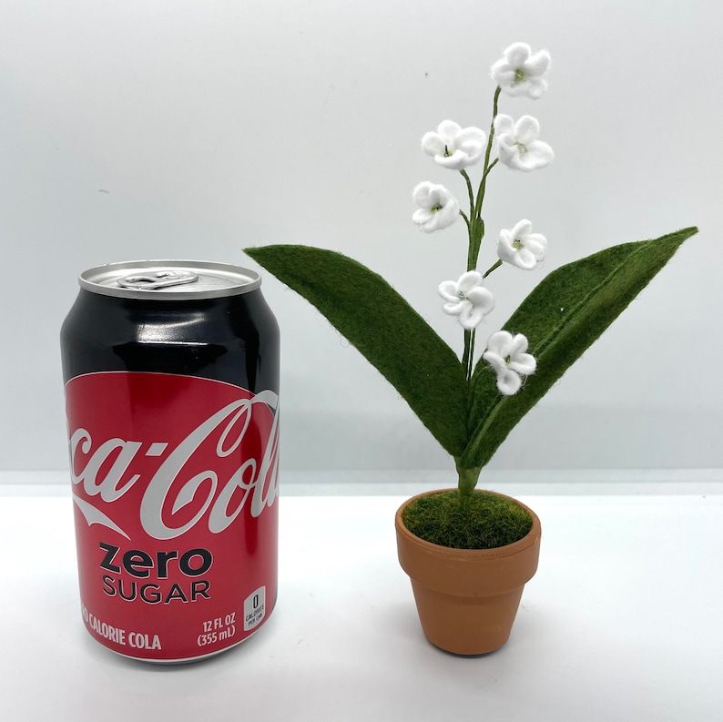 Peut inclure: Une petite plante en pot avec des fleurs blanches en feutre et des feuilles vertes en feutre. La plante est dans un pot en terre cuite brun et est pos&eacute;e sur une surface blanche &agrave; c&ocirc;t&eacute; d'une canette de Coca-Cola Zero Sugar.
