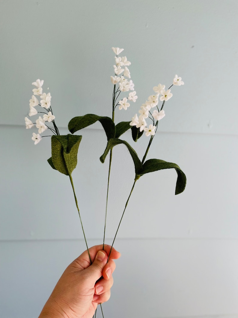 Peut inclure: Trois tiges de fleurs en feutre blanc avec des feuilles en feutre vert. Les fleurs sont dispos&eacute;es en bouquet et tenues dans une main.