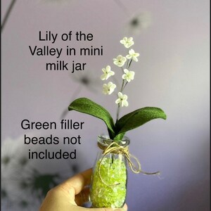 Peut inclure: Un petit arrangement floral de muguet blanc dans un mini pot &agrave; lait avec des billes de remplissage vertes. Le texte "Lily of the Valley in mini milk jar" et "Green filler beads not included" est visible sur l'image.