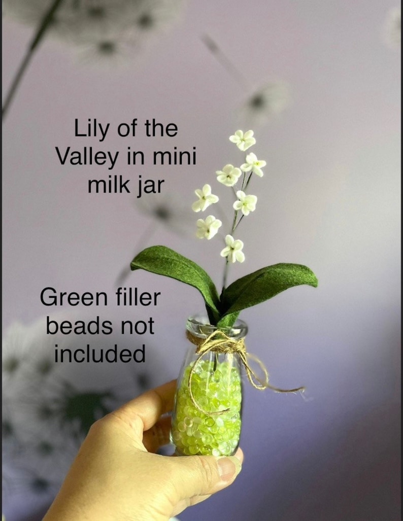 Peut inclure: Un petit arrangement floral de muguet blanc dans un mini pot &agrave; lait avec des billes de remplissage vertes. Le texte "Lily of the Valley in mini milk jar" et "Green filler beads not included" est visible sur l'image.