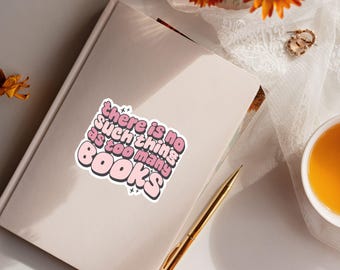 May include: A rectangular sticker featuring the text "there is no such thing as too many BOOKS" in a bold, pink, bubble-style font with a dark outline, placed on the cover of a closed, light-colored notebook next to a gold-toned pen.