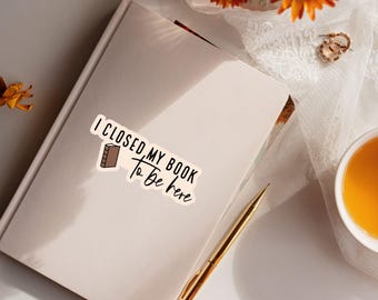 May include: A rectangular sticker with a cream background featuring the black text I CLOSED MY BOOK to be here and a small brown book icon. The sticker is placed on a closed off-white notebook next to a gold pen and decorative elements.