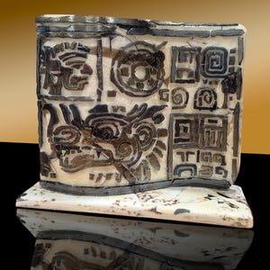 May include: A rectangular, off-white sculpture with intricate, dark brown carvings. The design features a stylized face, circular patterns, and geometric shapes. The sculpture sits on a matching base, reflecting the light.