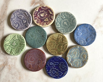May include: A collection of ten ceramic coasters in various colors, including green, blue, purple, and brown. Each coaster features a raised floral design. The coasters are round and appear to be handmade.