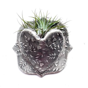 May include: A heart-shaped gray ceramic wall planter featuring an embossed leaf pattern across its surface. The planter holds a small green air plant with thin, spiky leaves protruding from the top opening of the heart-shaped vessel.